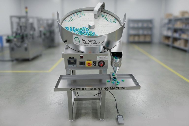 Capsule counting machine in a clean lab, blue and green capsules on the rotary pan and tray labeled Capsule Counting Machine (Adinath International).