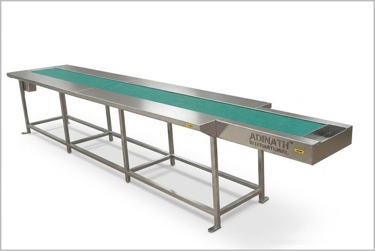 Long stainless steel conveyor with a green belt on a metal frame, branded ADINATH International, set against a white studio backdrop.